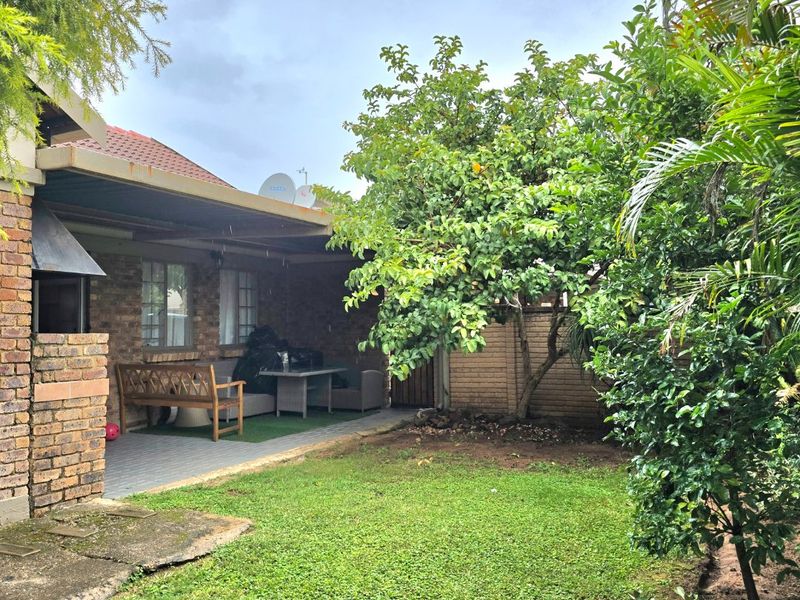 Rear garden area featuring a covered patio with a built-in braai, brick exterior walls, and established landscaping.
