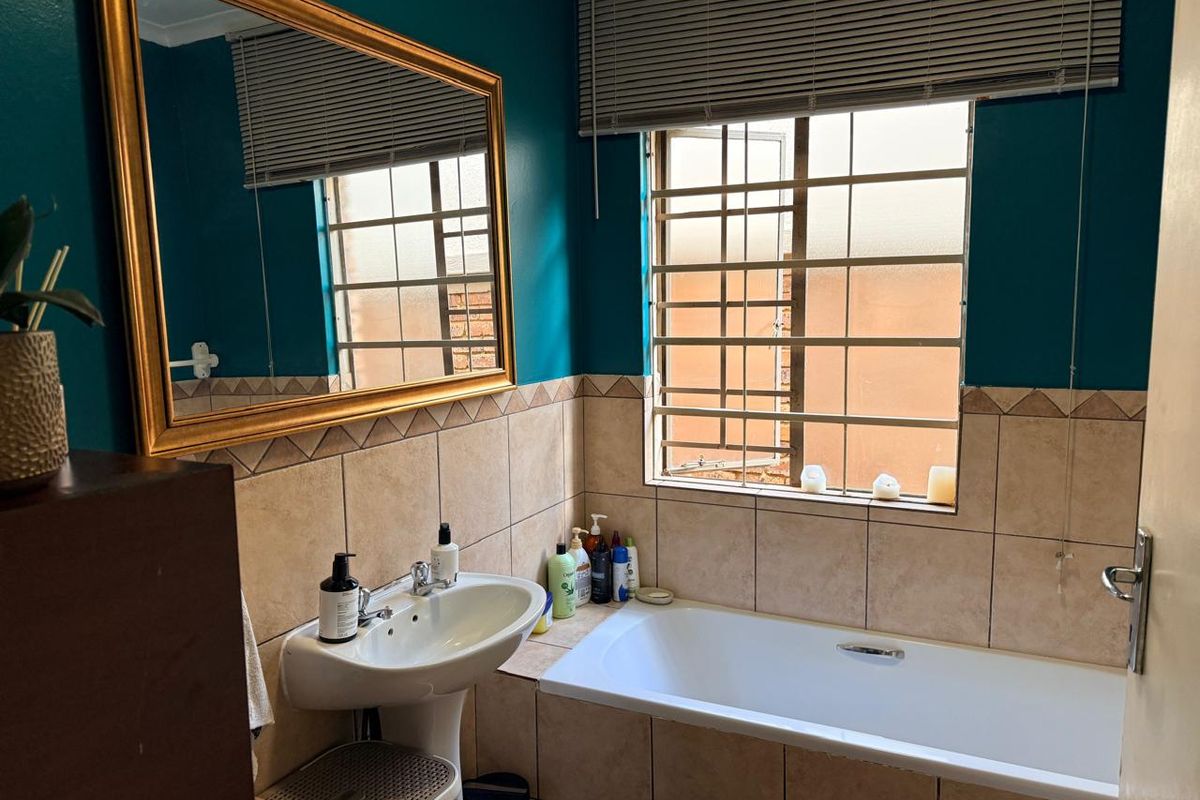 A functional bathroom featuring a pedestal basin, built-in bathtub, and tiled splashback, complemented by security bars on the window and a wall-mounted mirror.