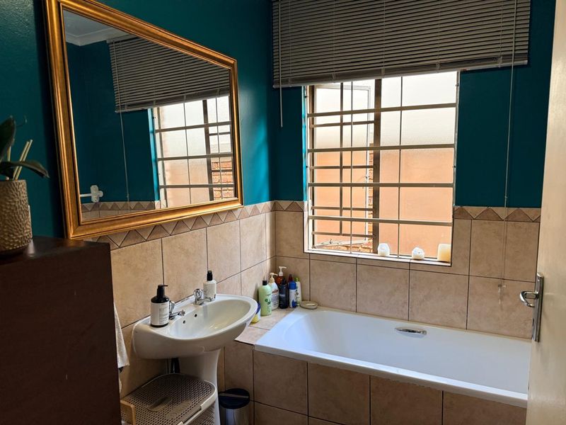 A functional bathroom featuring a pedestal basin, built-in bathtub, and tiled splashback, complemented by security bars on the window and a wall-mounted mirror.