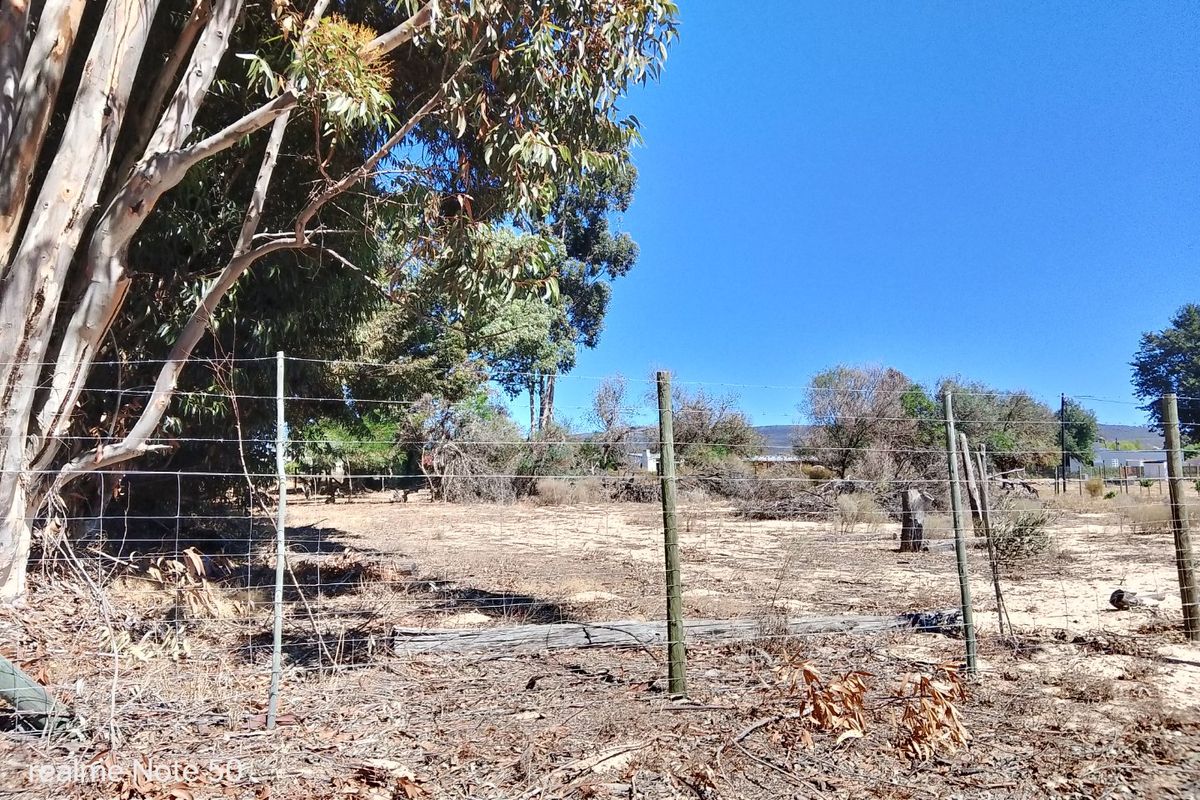 A view of a vacant residential plot featuring wire fencing, wooden posts, and mature trees on a dry, unimproved landscape.