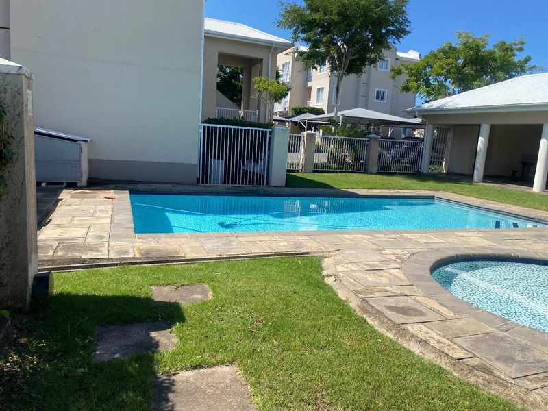 Outdoor pool area featuring a swimming pool with paved surround, lawn space, and access to a covered patio area within a secure complex.