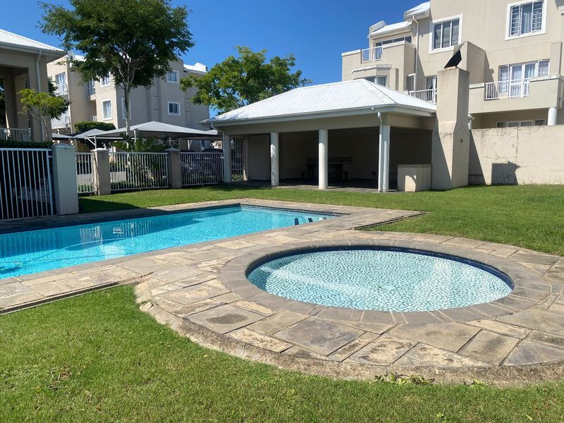 Communal outdoor area featuring a swimming pool, circular spa, and a covered patio with an outdoor fireplace, set within a landscaped garden.
