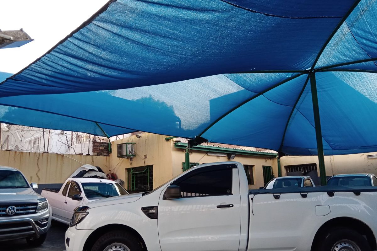 Commercial outdoor parking area featuring a large blue shade cloth structure, perimeter wall with security fencing, and external building infrastructure.