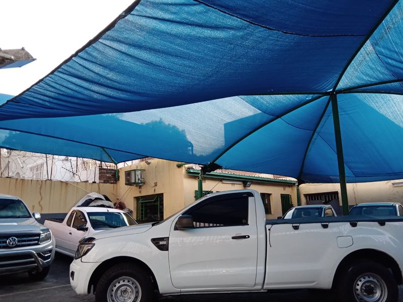 Commercial outdoor parking area featuring a large blue shade cloth structure, perimeter wall with security fencing, and external building infrastructure.