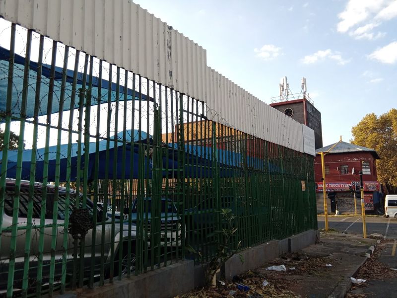 Street-level view of a commercial property featuring security fencing with barbed wire, corrugated metal cladding, and shaded parking areas.