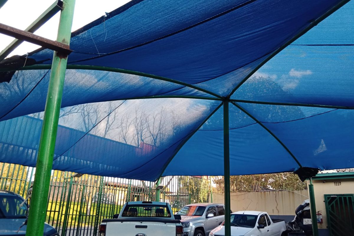 Outdoor parking area featuring a blue shade cloth canopy supported by metal poles and enclosed by security fencing.