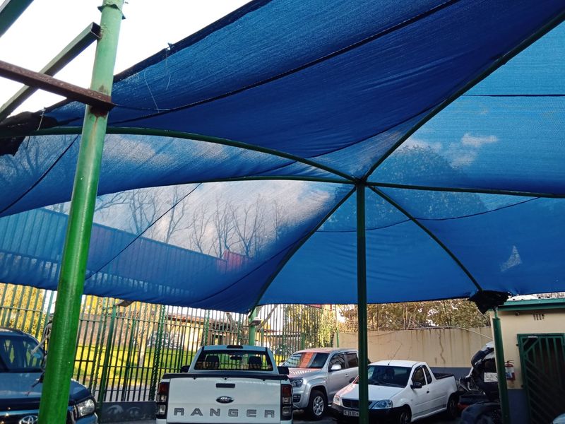 Outdoor parking area featuring a blue shade cloth canopy supported by metal poles and enclosed by security fencing.