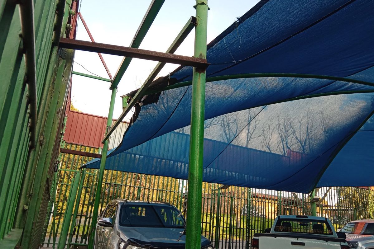 Outdoor parking area featuring a metal shade structure with damaged shade cloth and perimeter security fencing.