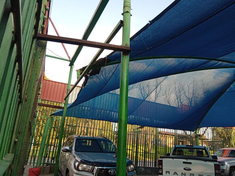 Outdoor parking area featuring a metal shade structure with damaged shade cloth and perimeter security fencing.