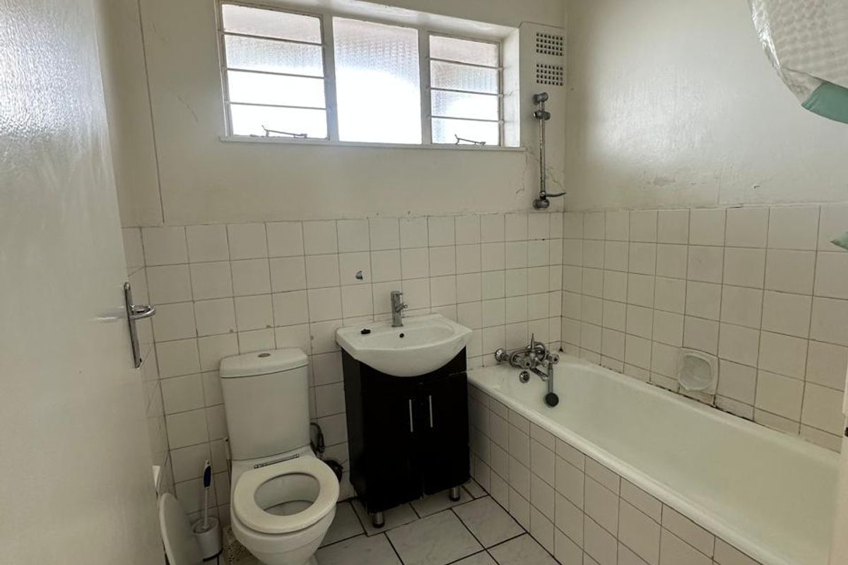 A dated bathroom featuring a standard bathtub, toilet, and vanity basin with white tiled walls and flooring, requiring cosmetic updates.