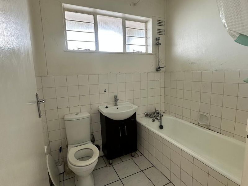 A dated bathroom featuring a standard bathtub, toilet, and vanity basin with white tiled walls and flooring, requiring cosmetic updates.