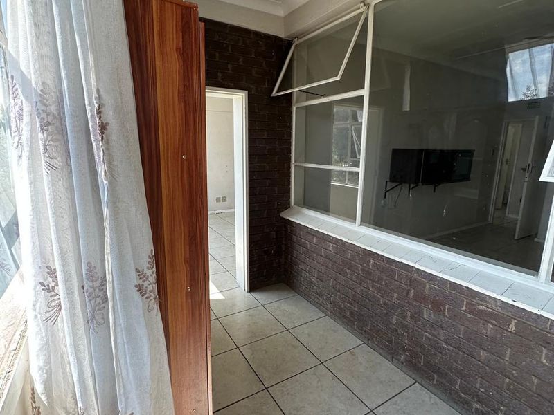 An enclosed balcony or sunroom featuring tiled flooring, exposed brick walls, and large casement windows, offering a versatile space for potential renovation.