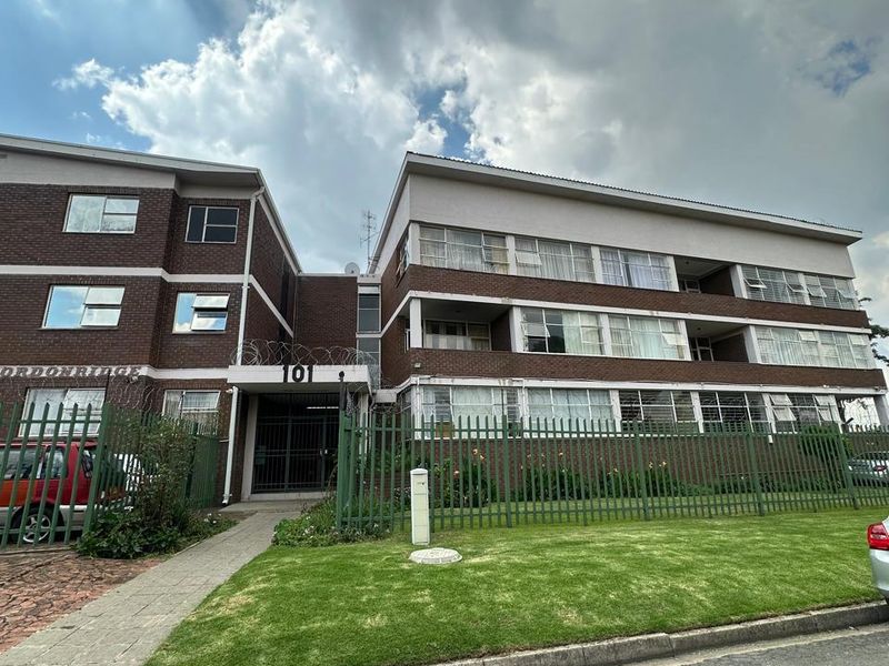 Front exterior view of a multi-story brick apartment building featuring secure perimeter fencing, a gated entrance, and a well-maintained front lawn.
