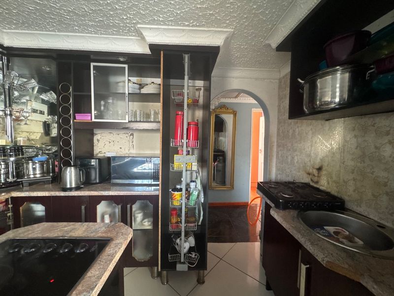A dated kitchen featuring dark cabinetry, dual hobs, and a pull-out pantry unit, leading through an archway to an adjacent room.