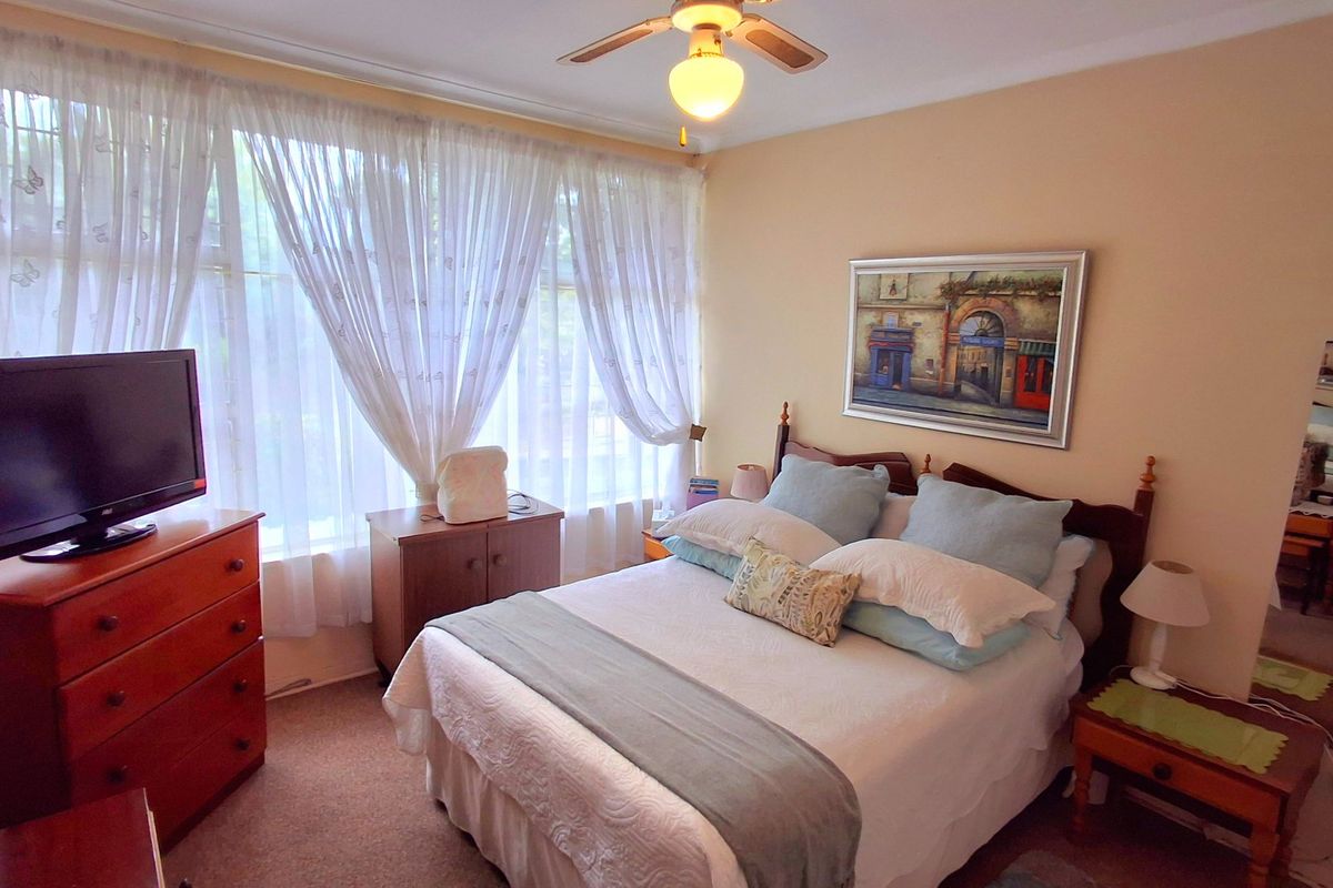 A cozy bedroom featuring a ceiling fan, large window with natural light, and carpeted flooring.