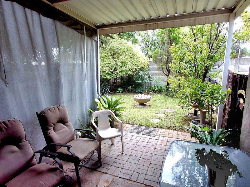 A covered patio area featuring paved flooring and a corrugated roof, overlooking a private garden with established greenery and a boundary wall.