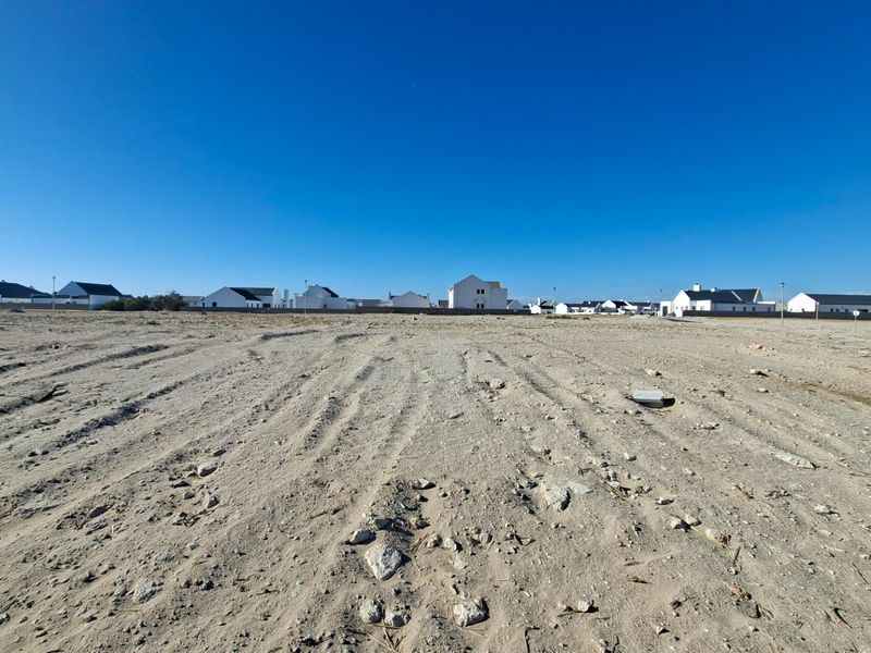A wide-angle view of a vacant residential plot featuring sandy terrain with surrounding established residential properties in the background.