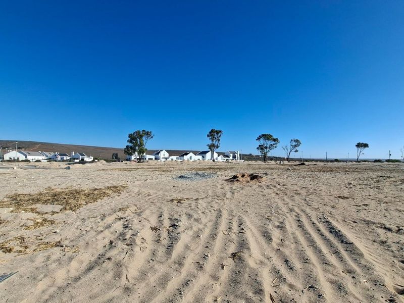 A wide-angle view of a vacant residential plot featuring sandy terrain with surrounding neighborhood development in the distance.