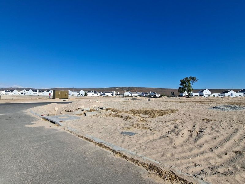 A vacant residential plot featuring an asphalt road frontage, concrete curbing, and an installed utility box, situated in a developing residential estate.