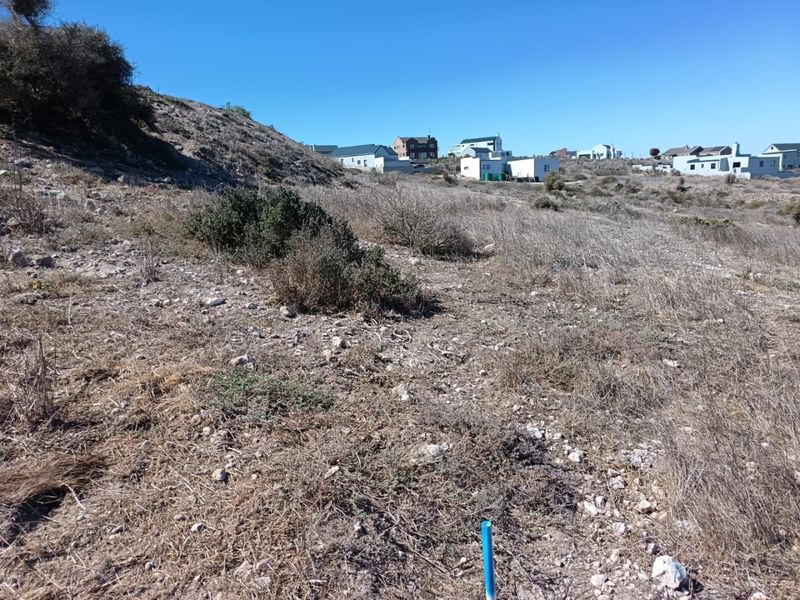 A view of a vacant residential plot featuring sloped terrain and natural vegetation, situated in a developed neighborhood.