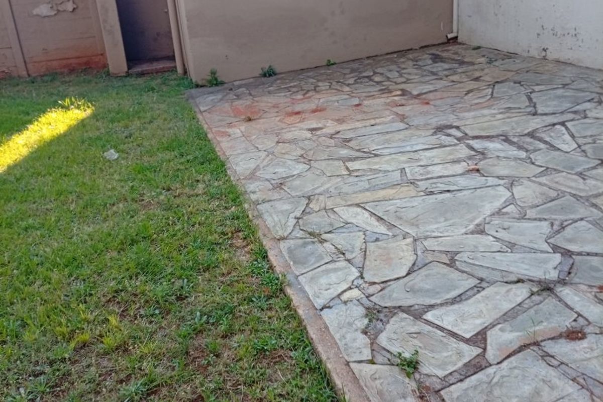 A rear exterior area featuring a stone-paved patio, a small lawn, and a boundary wall requiring maintenance.