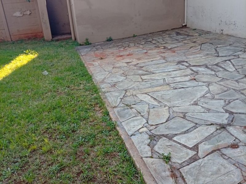 A rear exterior area featuring a stone-paved patio, a small lawn, and a boundary wall requiring maintenance.