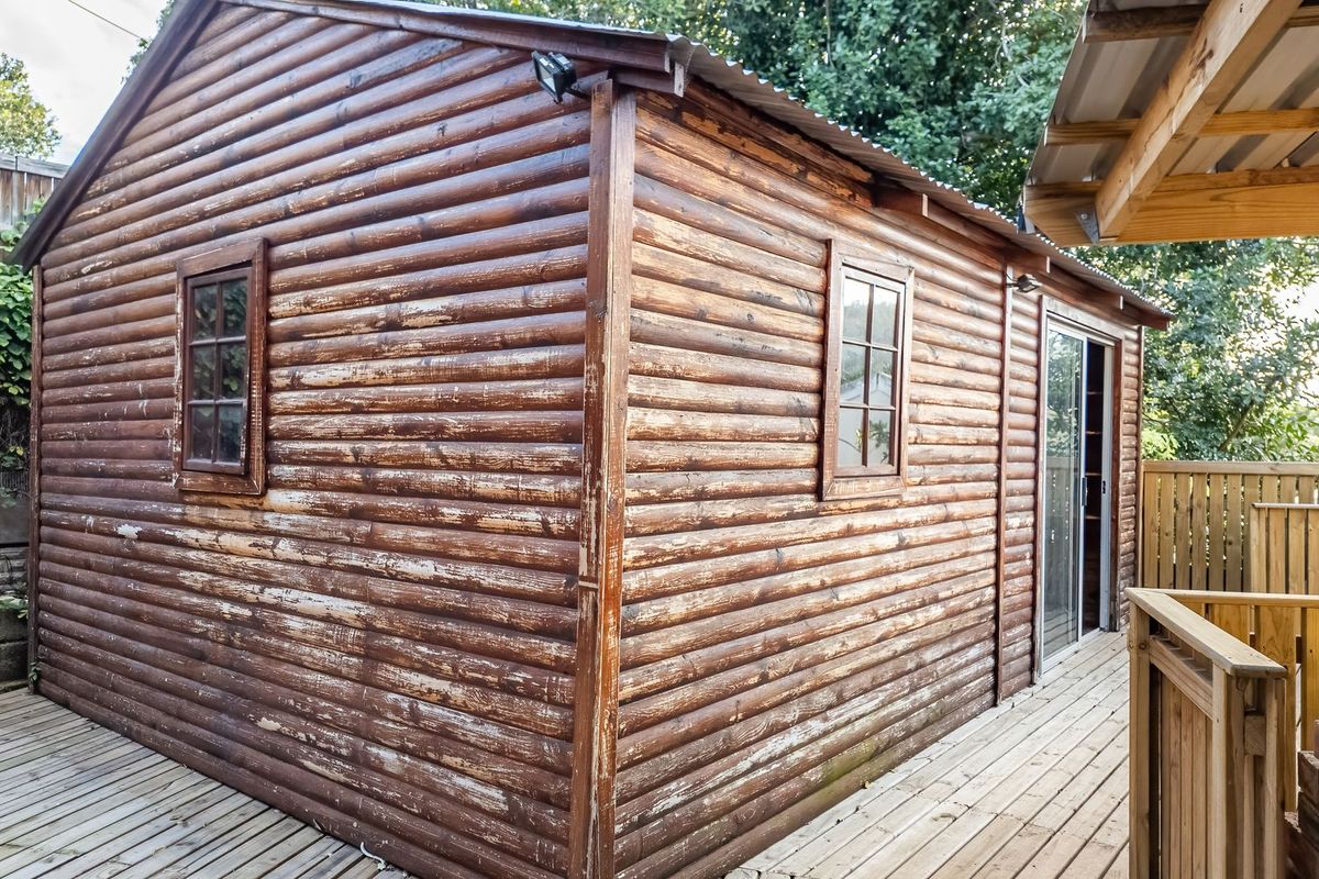 A detached log cabin structure featuring timber decking, sliding glass doors, and timber-framed windows, showing signs of exterior wood weathering.
