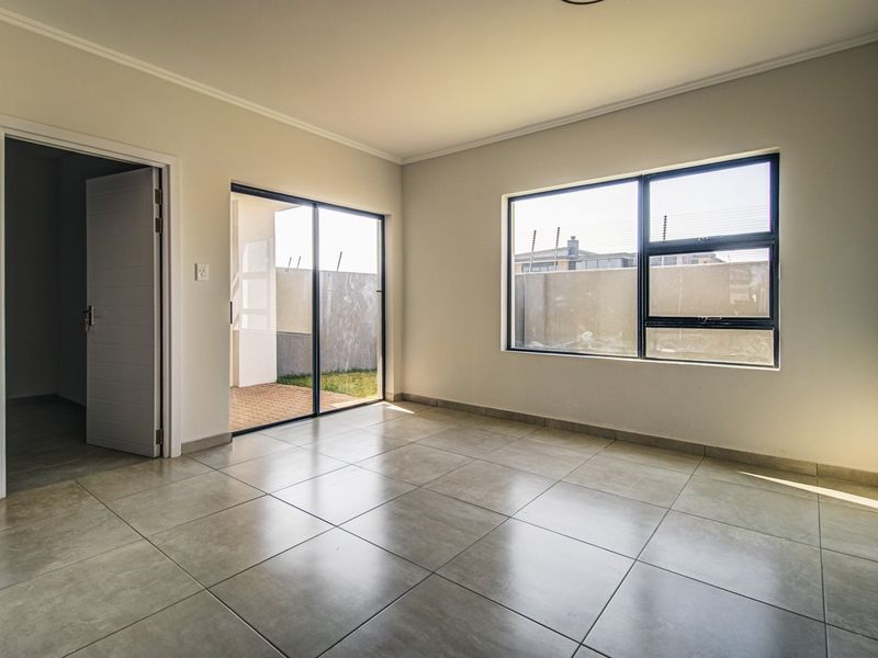 A bright, empty living room featuring neutral tiled flooring, large aluminium-framed windows, and a sliding glass door providing access to the outdoor area.