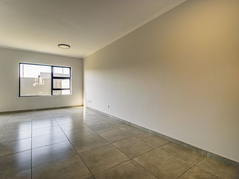 A bright, empty living room featuring neutral tiled flooring, clean walls, and a large aluminium-framed window providing natural light.