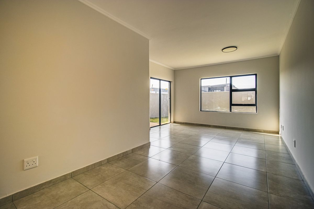 A bright, empty living area featuring neutral tiled flooring, modern aluminium window frames, and a sliding door leading to the exterior.