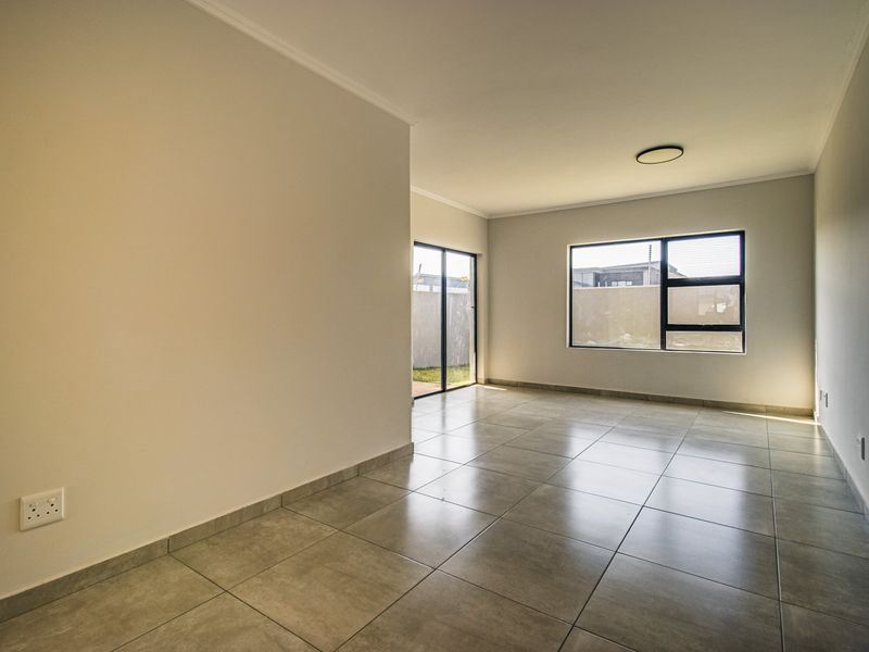 A bright, empty living area featuring neutral tiled flooring, modern aluminium window frames, and a sliding door leading to the exterior.