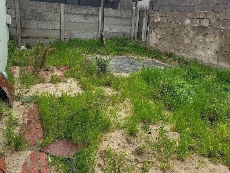 A rear outdoor area featuring a concrete boundary wall, brick paving, and a concrete slab, currently requiring significant landscaping and maintenance.