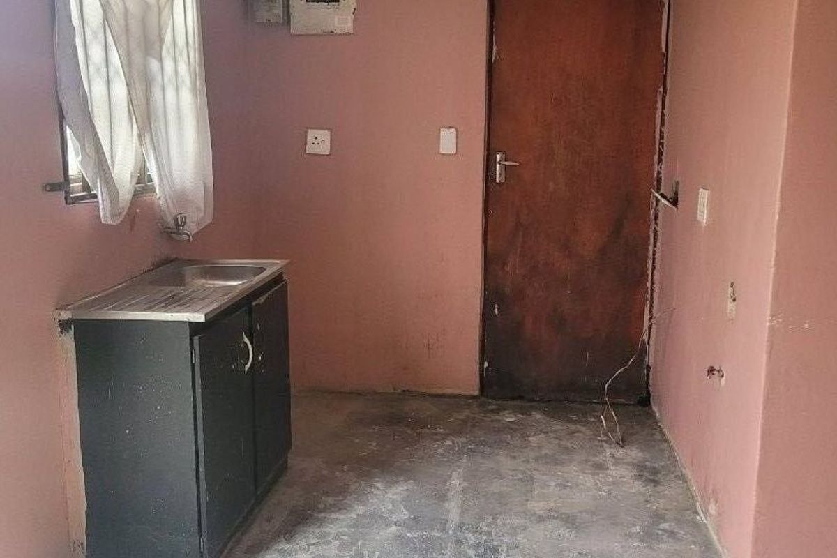 A basic, empty kitchen area featuring a single basin sink, base cabinetry, and an electrical distribution board, requiring significant cosmetic updates and flooring repairs.