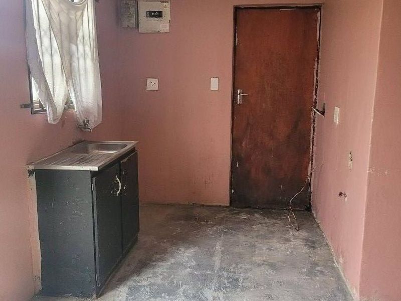 A basic, empty kitchen area featuring a single basin sink, base cabinetry, and an electrical distribution board, requiring significant cosmetic updates and flooring repairs.