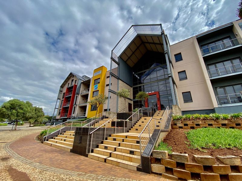 Modern commercial building facade featuring a prominent glass entrance, steel architectural elements, and tiered stone landscaping with paved pedestrian access.