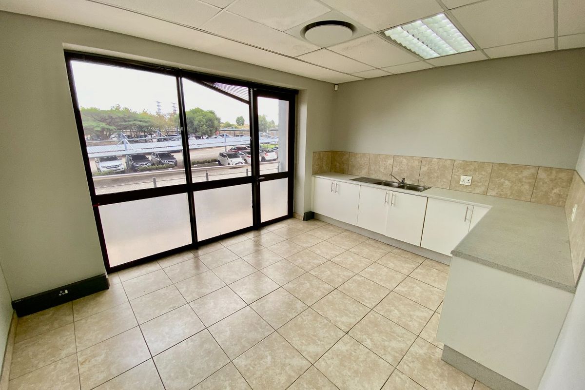A functional, empty commercial kitchenette featuring tiled flooring, built-in cabinetry with a stainless steel sink, and large windows providing natural light.