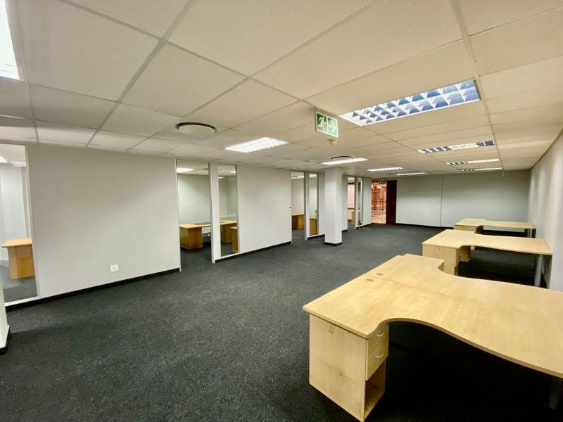 Open-plan commercial office space featuring glass-partitioned private offices, suspended ceilings with recessed lighting, and dark carpeting.