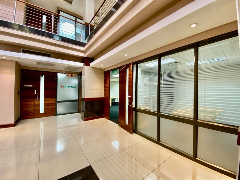 A modern commercial hallway featuring tiled floors, glass-partitioned office spaces, and a mezzanine level with a stainless steel balustrade.
