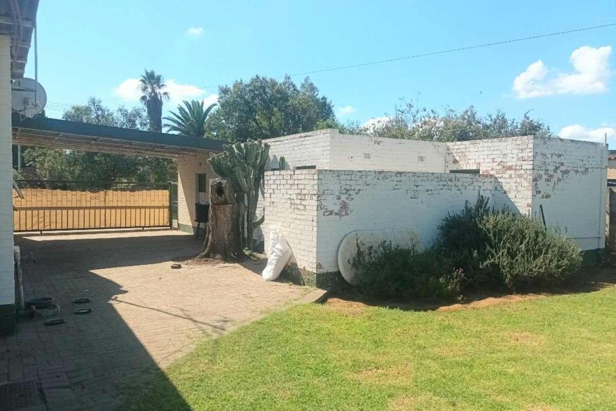 Rear exterior view of the property featuring a paved driveway, carport, metal gate, and a brick boundary wall with a lawn area.