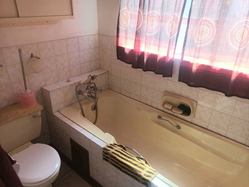 A dated bathroom featuring a built-in bathtub with wall-mounted mixer, tiled walls, and a standard toilet, offering potential for modernization.