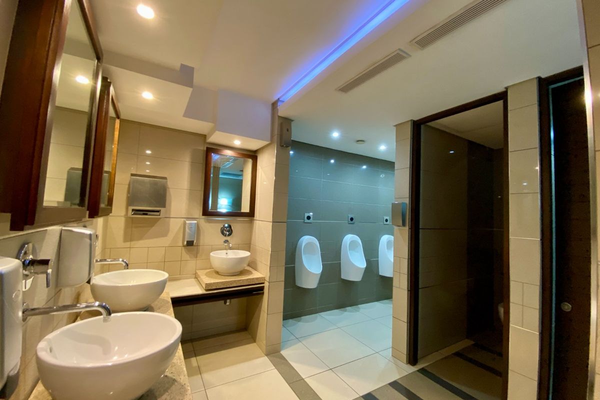 A modern commercial restroom featuring multiple vessel sinks on stone vanity tops and a dedicated urinal area with tiled finishes and ambient lighting.