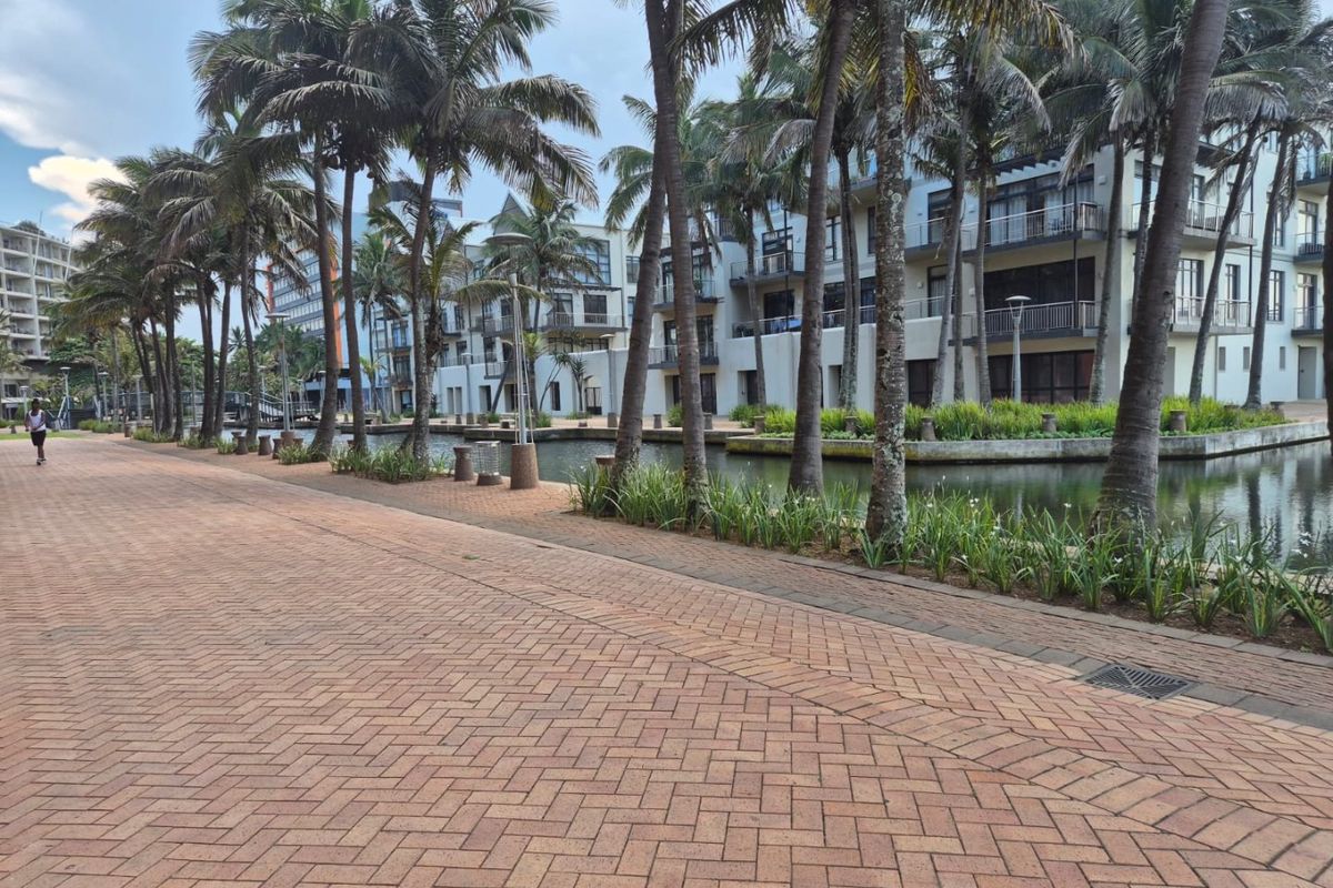 A scenic paved walkway alongside a tranquil canal, featuring mature palm trees and views of modern apartment building facades with balconies.