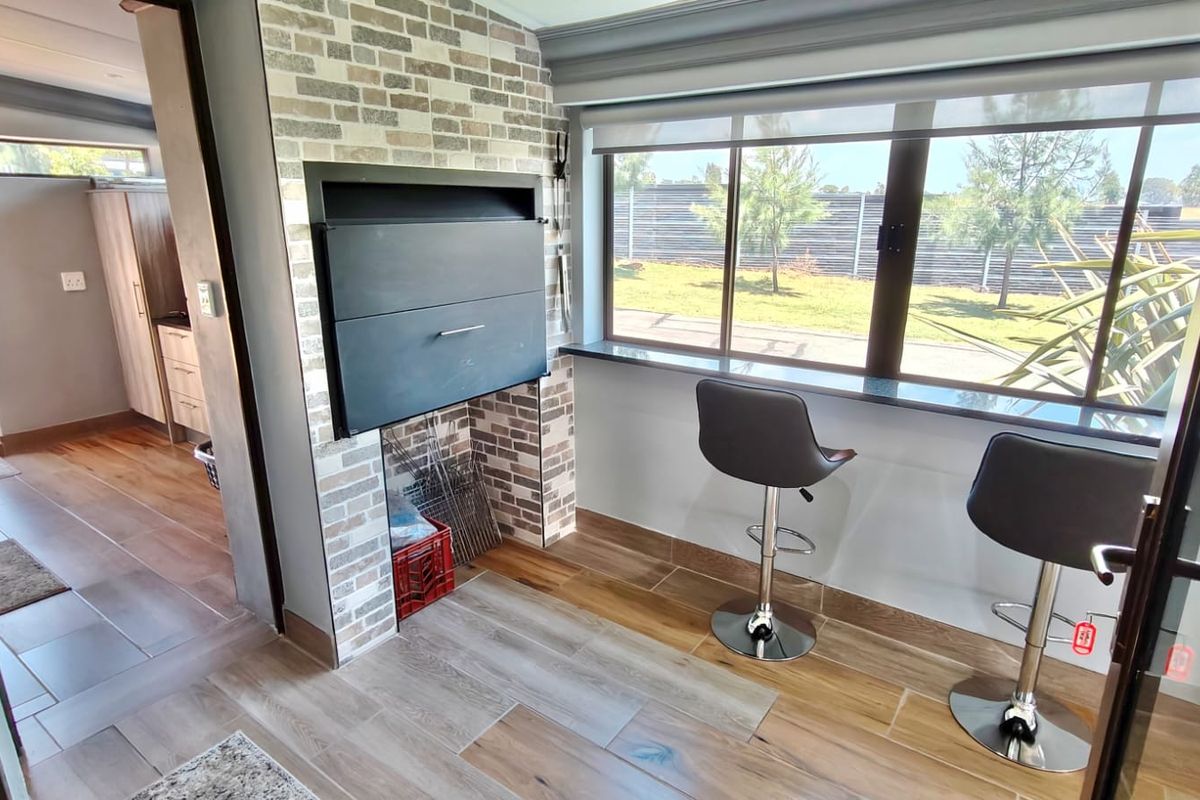 A bright living area featuring a built-in braai with a brick surround, large windows, and modern tiled flooring.