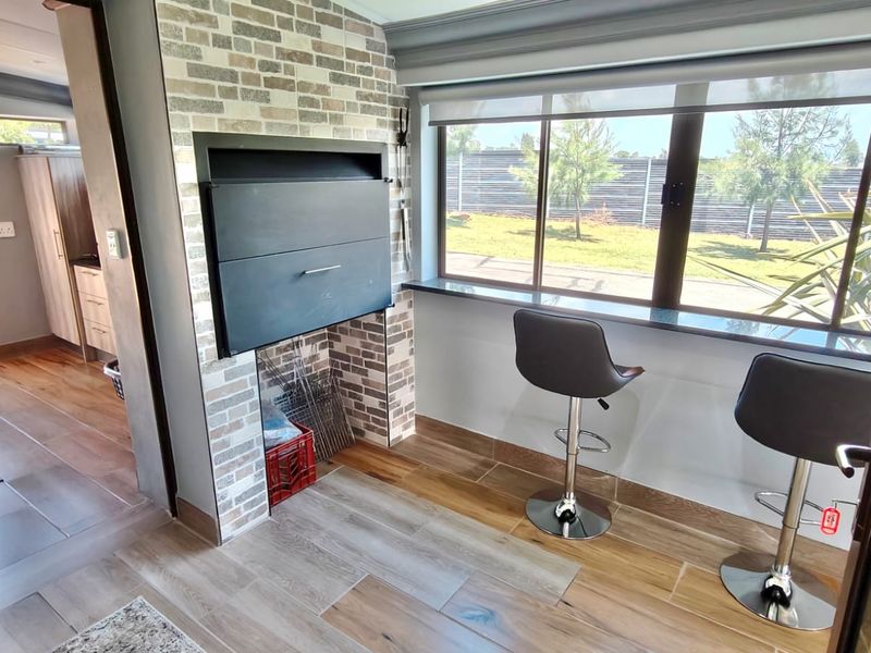A bright living area featuring a built-in braai with a brick surround, large windows, and modern tiled flooring.