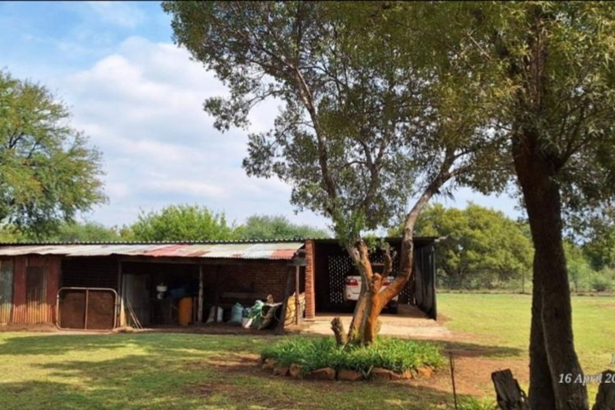 A rustic farm storage shed and carport structure featuring corrugated iron roofing and brick construction set within a large open garden area.