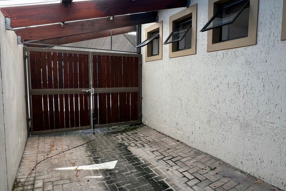 A paved rear exterior area featuring a timber gate, pergola structure, and textured plaster walls with top-hung windows.