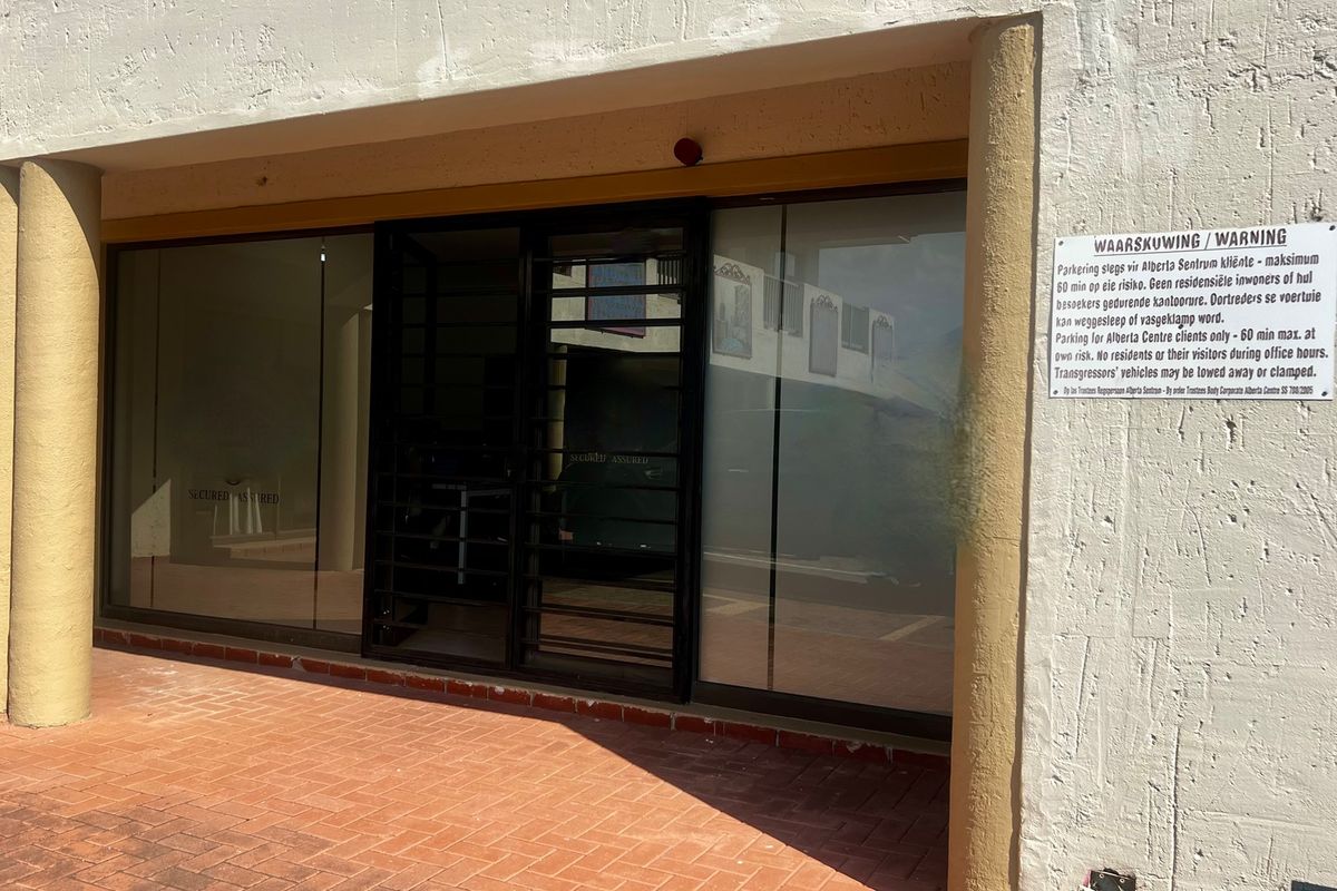 Commercial storefront entrance featuring a glass facade, security gate, and brick paved walkway, with signage indicating parking restrictions.