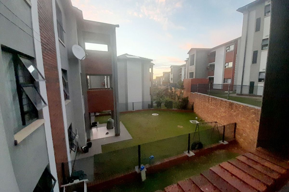 An elevated view of the apartment complex grounds, featuring a shared lawn area, brick boundary walls, and surrounding residential building facades.