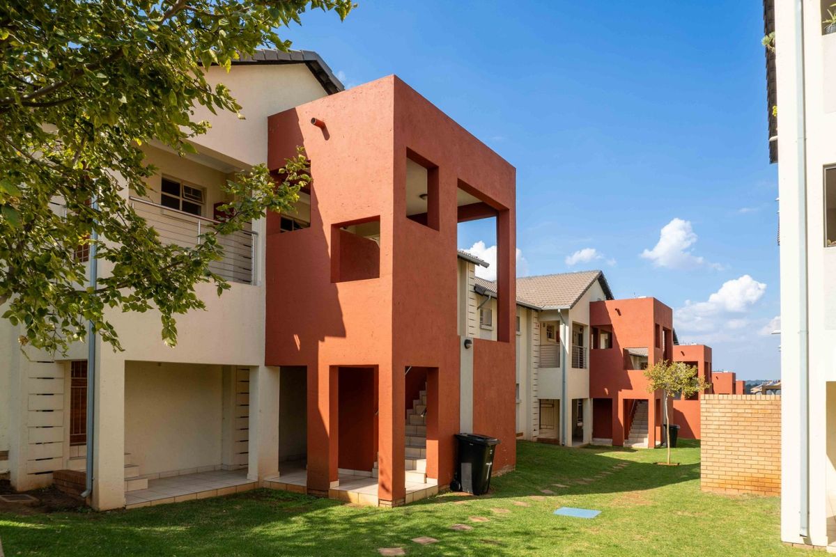 Exterior view of a modern apartment complex featuring red-toned architectural facades, private patios, balconies, and well-maintained communal garden spaces.