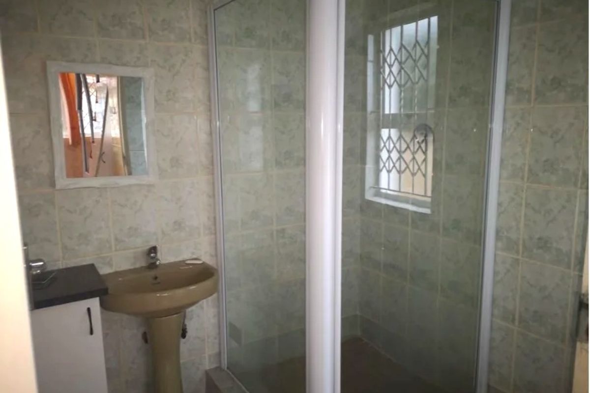A functional bathroom featuring a corner shower enclosure, pedestal basin, and full-height wall tiling with security bars on the window.